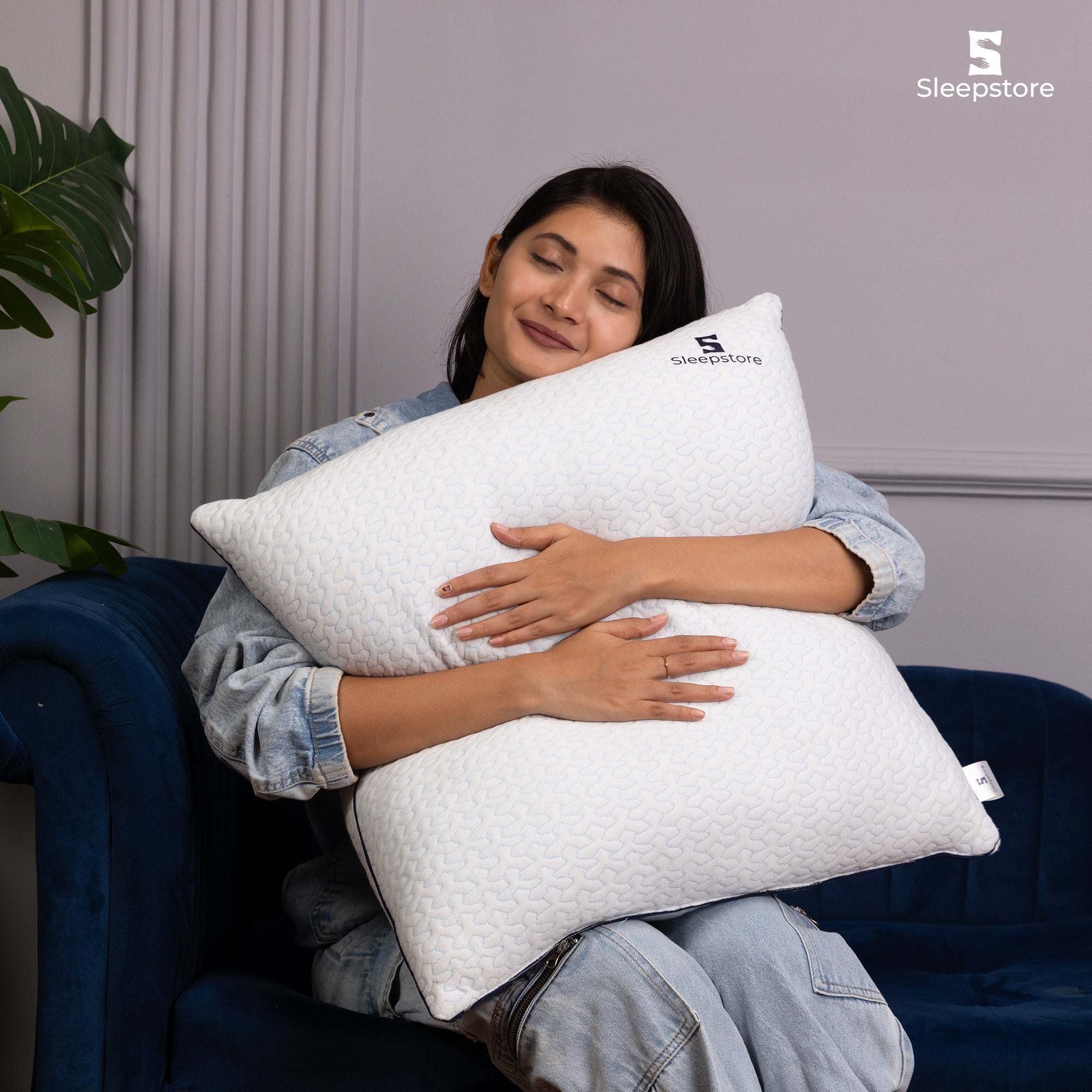 Woman in denim outfit hugging a white Sleepstore pillow while sitting on a blue sofa.