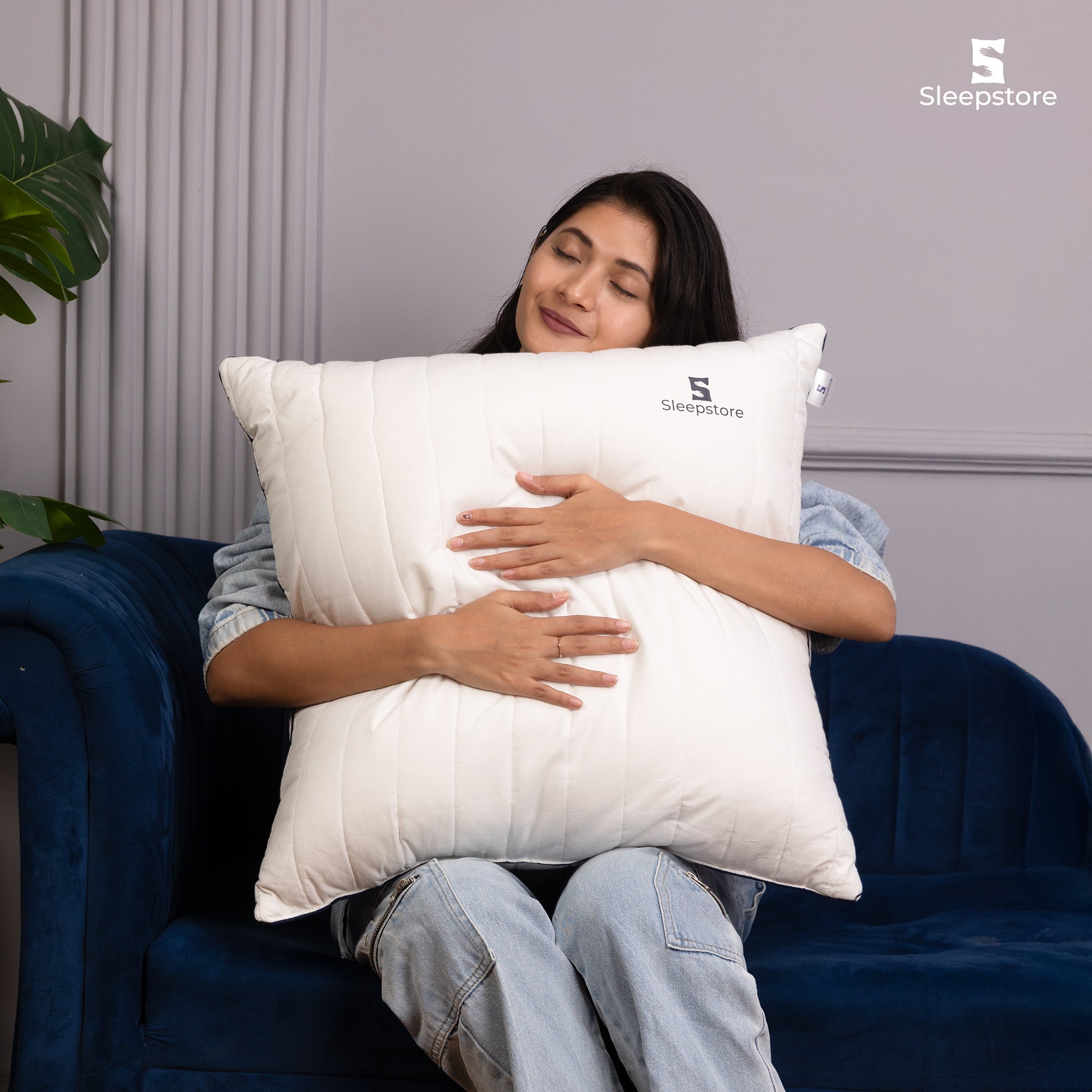 Woman sitting on sofa hugging a large white Sleepstore cushion with a relaxed smile.
