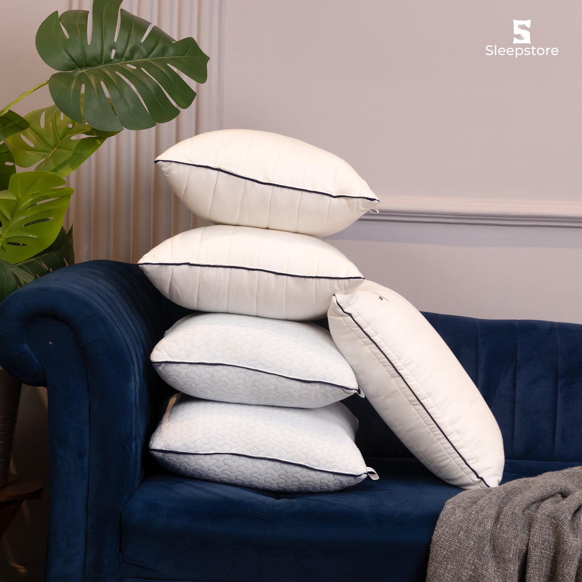 Stack of white Sleepstore pillows with black piping placed on a blue velvet sofa.
