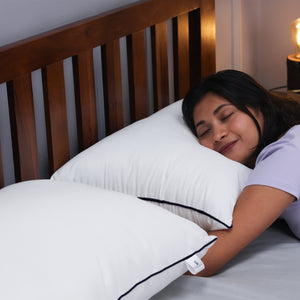 Woman peacefully hugging white Sleepstore pillow with black piping, lying on bed for cozy rest.