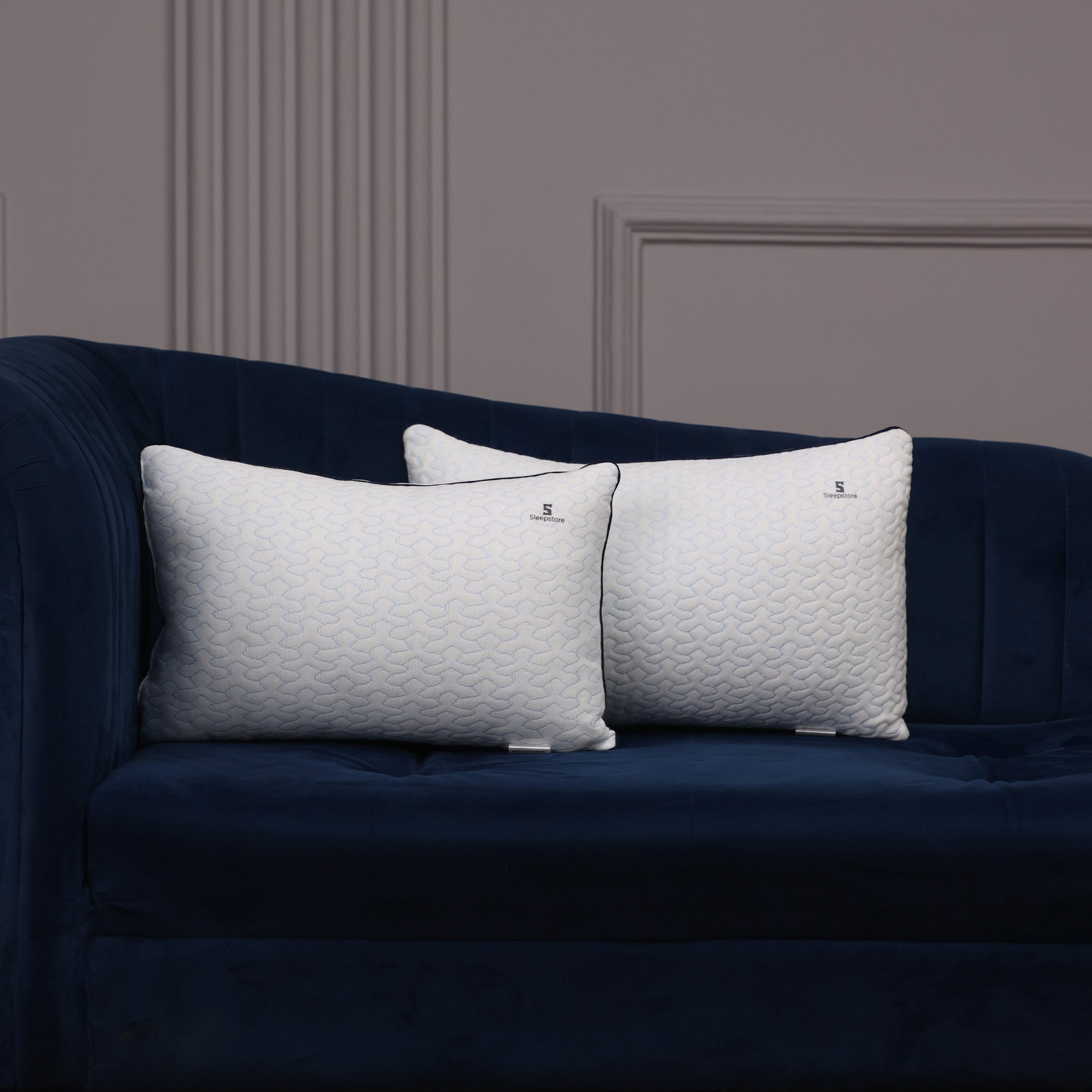 Two white Sleepstore pillows placed on a dark blue velvet sofa against a gray wall.