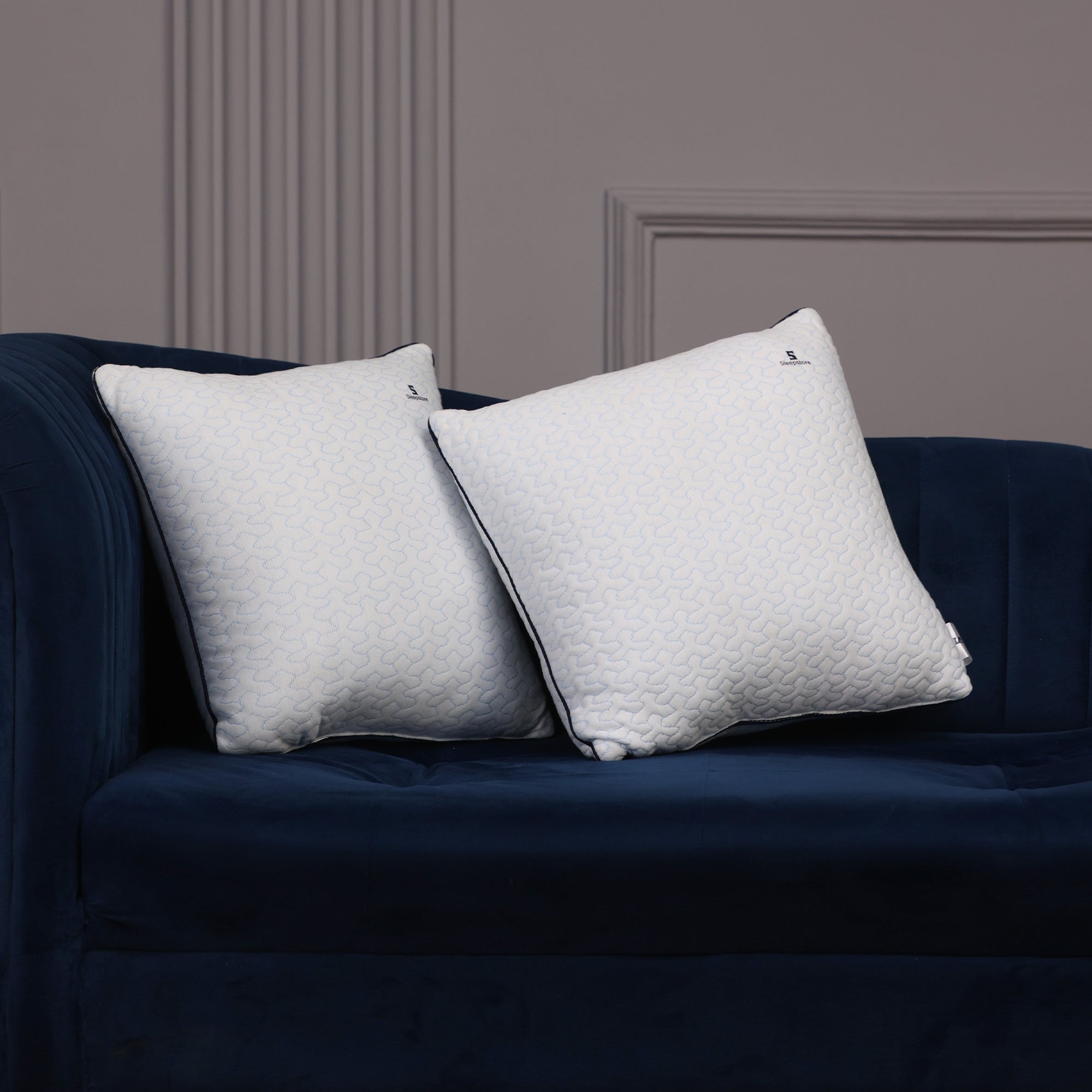 Two white square Sleepstore pillows resting on a dark blue velvet sofa against a gray wall.