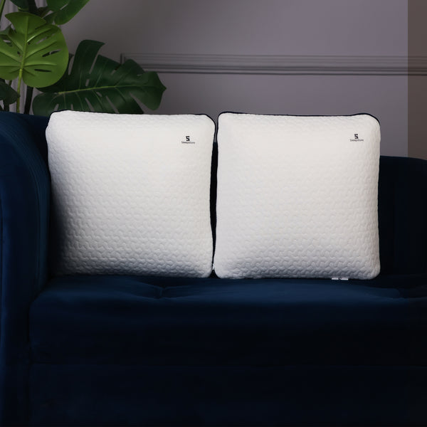 Two white square Sleepstore pillows with textured covers placed on a dark blue sofa beside green indoor plants.