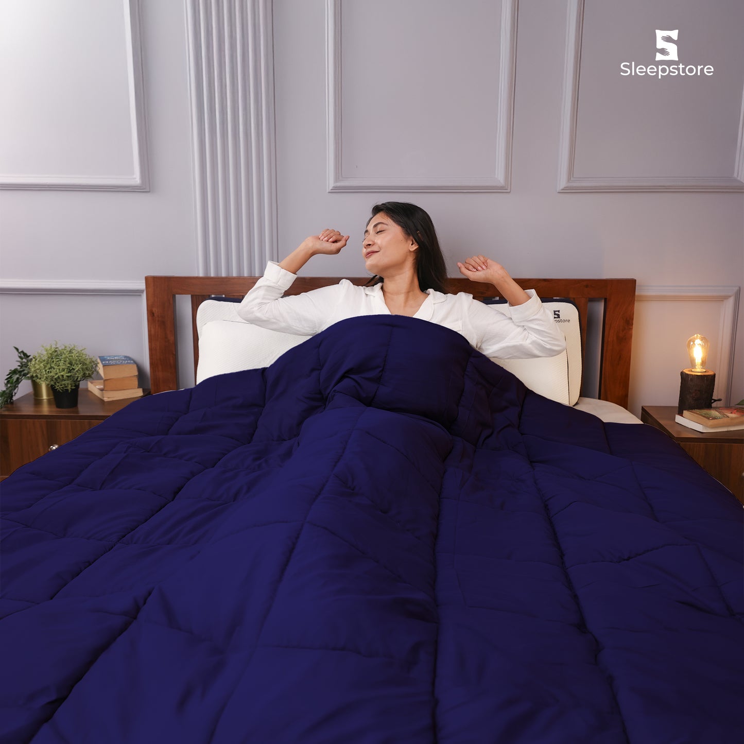 Woman stretching in bed under a cozy navy blue comforter, looking relaxed and happy.