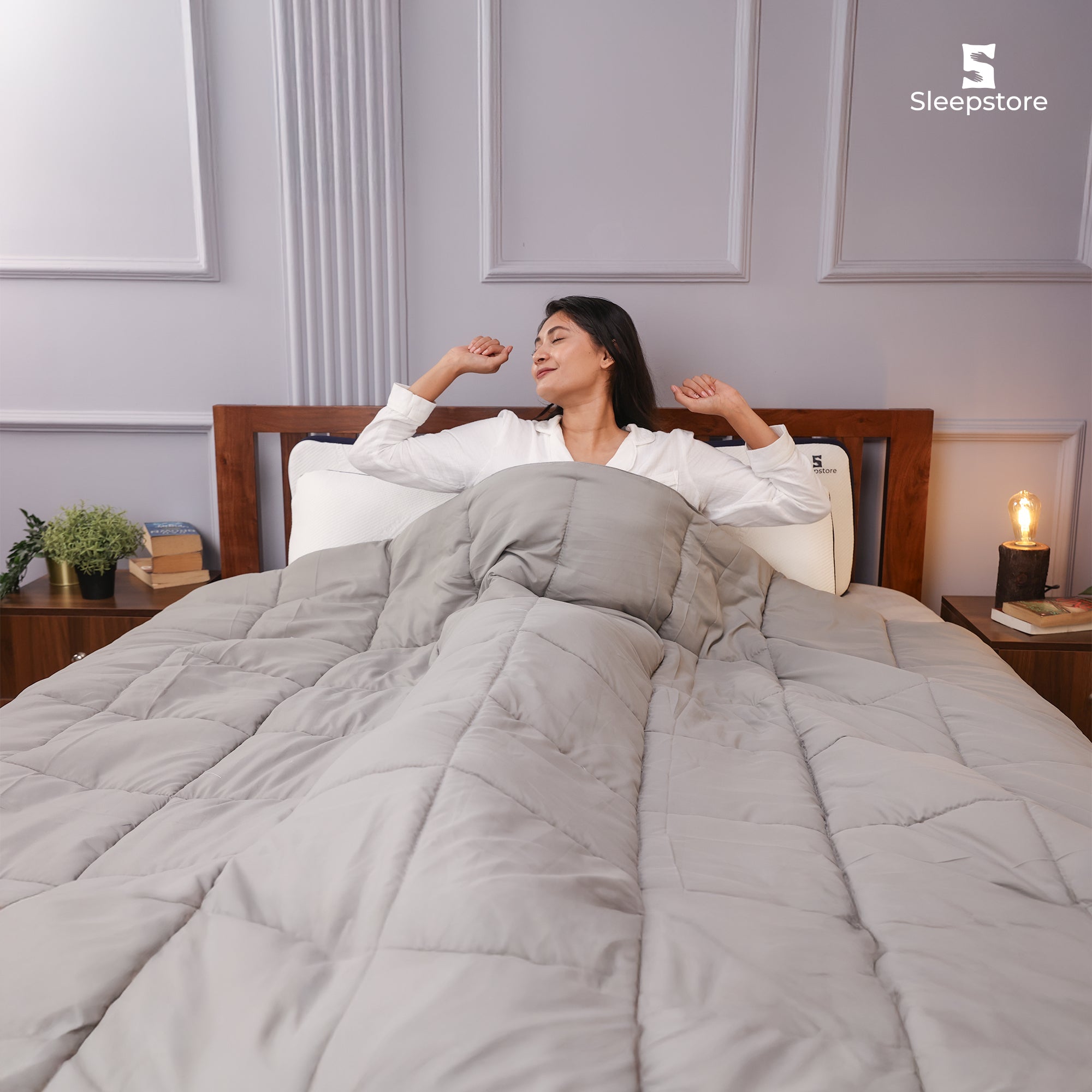 Woman stretching in bed under a grey quilted blanket with cozy bedroom setup.