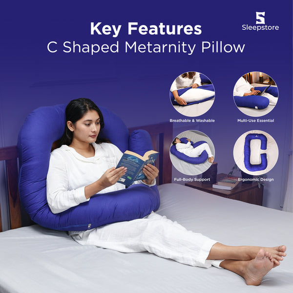 Woman reading a book while using a blue C-shaped maternity pillow on bed.